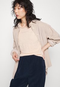 Camicia di corduroy beige sopra una t-shirt beige chiaro, abbinata a pantaloni navy texture. La camicia presenta bottoni prominenti e una vestibilità rilassata.