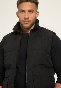 Gilet imbottito nero con colletto alto, zip frontale e due tasche grandi. Tessuto testurizzato con una finitura liscia e dettagli minimi.