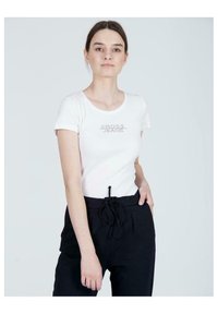 Biała koszulka z krótkim rękawem, okrągłym dekoltem i logiem "CROSS JEANS" wyłożonym kryształkami na piersi, zestawiona z czarnymi spodniami z troczkami.