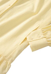 Żółta koszula z długim rękawem wykonana z lekkiego materiału. Posiada zapinane mankiety oraz zdobienia w formie marszczenia u dołu rękawa. Gładka tekstura.