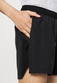 Pantalones cortos deportivos negros con una tela texturizada, cintura elástica y un bolsillo lateral con cierre de cremallera. De corta longitud con aberturas laterales.