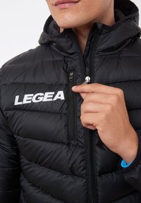 Svart puffjacka med quiltad textur, dragkedjestängning och huva. Har en vit broderad logotyp på bröstet. Händer som justerar dragkedjan.