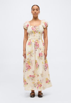 Femme debout, droite, portant une longue robe crème avec des motifs floraux roses et jaunes, des manches courtes bouffantes, associée à des chaussures plates foncées.