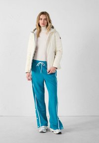 Chaqueta beige acolchada con capucha, sobre un cuello de tortuga blanco de canalé, combinada con pantalones deportivos en color teal con rayas laterales blancas y botones a presión. Zapatillas blancas.