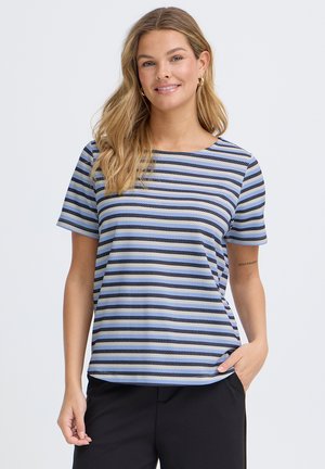 Lächelnde Frau mit langen blonden Haaren trägt ein blau-schwarz gestreiftes kurzärmeliges Shirt und schwarze Hosen, mit einer Hand in der Tasche.