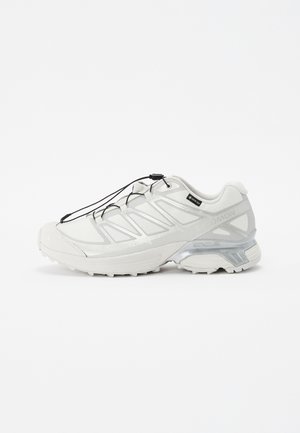 Sapato atlético branco com parte superior em malha texturizada, detalhes refletivos e sola amortecida. Apresenta atacadores elásticos e detalhes em cinza.