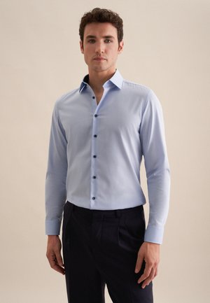 Homme debout portant une chemise à manches longues bleu clair avec des boutons noirs et un pantalon noir sur un fond beige uni.