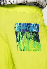 Limegrønne bukser med en stor lomme med et flerfarvet abstrakt mønster og lyseblå branding. Glat tekstur med en afslappet pasform.