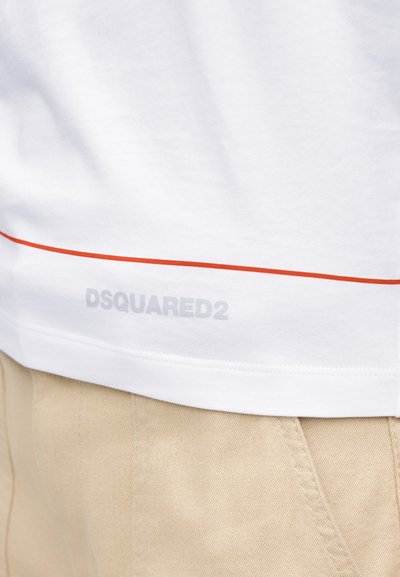 Λευκό τελείωμα πουκάμισου με κόκκινη οριζόντια γραμμή και αχνό λογότυπο "DSQUARED2" πάνω από την τσέπη μπεζ παντελονιού.