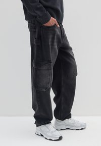 Zwarte denim cargobroek met een losse pasvorm, voorzien van meerdere zijzakken en een verweerde textuur. Gecombineerd met witte sportschoenen.