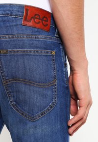 Blå denimjeans med en brun läderpatch med "Lee" på midjan, med kontrasterande sömmar, en böjd bakficka och en texturerad finish.