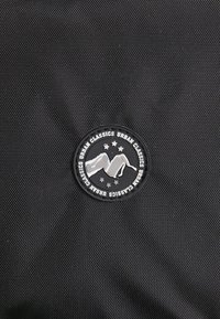 Tissu noir texturé arborant un logo circulaire noir et argent avec un design de montagne et le texte "Urban Classics".
