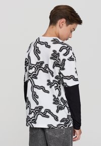 Nastolatek stoi bokiem, nosząc białą koszulkę z czarnymi wzorami przypominającymi błyskawice oraz długie czarne rękawy pod spodem.