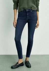 Tamno plave uske traperice s visokim strukom i glatkom teksturom. Uparene s crnim mokasinama i rebrastim zelenim puloverom.