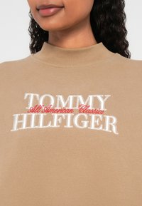 Μπεζ φούτερ με ψηλό λαιμό, με κεντημένο λογότυπο «TOMMY HILFIGER» σε λευκό και «All American Classics» σε κόκκινο στο ύψος του στήθους.