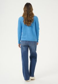 Modrý pletený svetr s dlouhými rukávy, zkombinovaný s volnými modrými džíny. Zadní pohled ukazuje žebrované manžety a pas.