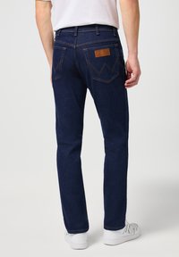 Jeans de mezclilla azul oscuro con corte recto, que presentan un parche de cuero marrón en la parte trasera y costuras amarillas en los bolsillos. Textura suave.