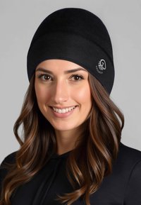 Czarna dopasowana czapka typu beanie wykonana z miękkiego materiału, z przeszyciem na górze i białym logo po boku, odpowiednia do noszenia na zewnątrz.