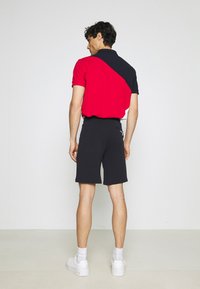 Polo da uomo in rosso con accenti neri, maniche corte e colletto, abbinato a pantaloni corti neri e sneakers bianche, visto da dietro.