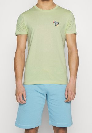 Mand iført en lys grøn kortærmet T-shirt med et lille zebra-patch og lyseblå knæ-lange shorts.