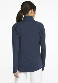 Maglia sportiva a maniche lunghe di colore blu navy con colletto alto, maniche raglan e orlo curvo. Realizzata in un tessuto liscio ed elasticizzato. Senza hardware visibile.