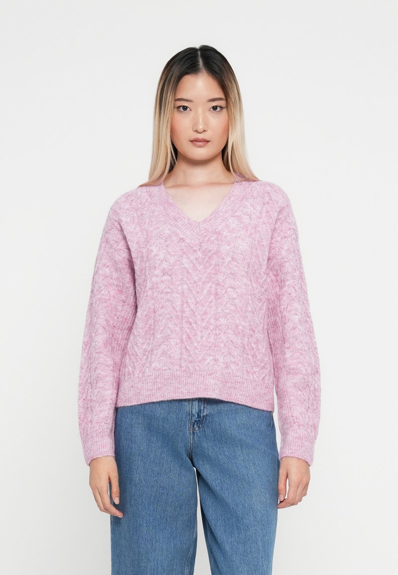 Rosa gestrickter Pullover mit V-Ausschnitt, der ein strukturiertes Muster und eine lockere Passform aufweist, kombiniert mit hellblauen Jeans.