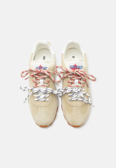 Μπεζ σουέντ αθλητικά παπούτσια με λευκά εσωτερικά, κόκκινο και λευκό λογότυπο μάρκας στη γλώσσα, και μη ταιριαστά κορδόνια — κόκκινα και λευκά στην κορυφή, μαύρα και λευκά από κάτω.