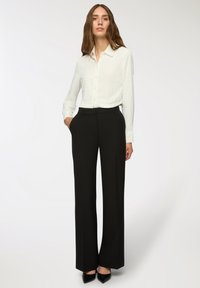 Camicia bianca a maniche lunghe con pois, abbinata a pantaloni neri a vita alta, completata da décolleté nere con punta. Look classico e su misura.