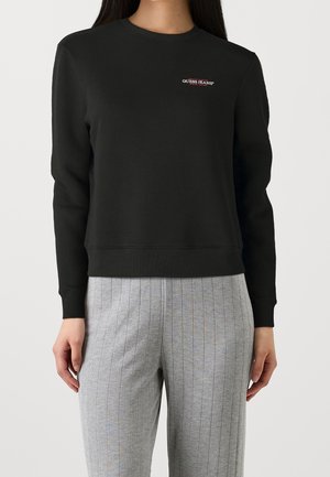 Nainen pukeutuneena mustaan Guess Jeans -collegepaitaan, jossa on pieni rintalogo, ja vaaleanharmaisiin, ohuella raidoituksella varustettuihin housuihin, seisoo yksinkertaista vaaleaa taustaa vasten.