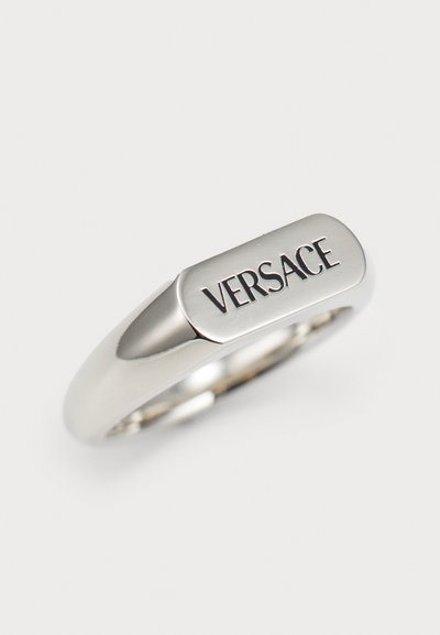 Zilveren ring met een glad, afgerond ontwerp en een rechthoekige bovenkant, gegraveerd met het woord "VERSACE" in vette, zwarte letters.