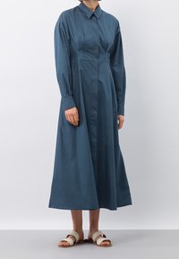 Abito a maniche lunghe e colletto in cotone di un profondo verde teal. Presenta un corpetto aderente, pieghe e una gonna svasata. Indossato con sandali piatti di colore chiaro.