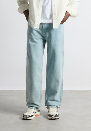 Homme portant un jean large bleu clair, des baskets blanches avec des détails noirs, un t-shirt blanc et une chemise écrue boutonnée, les mains dans les poches.