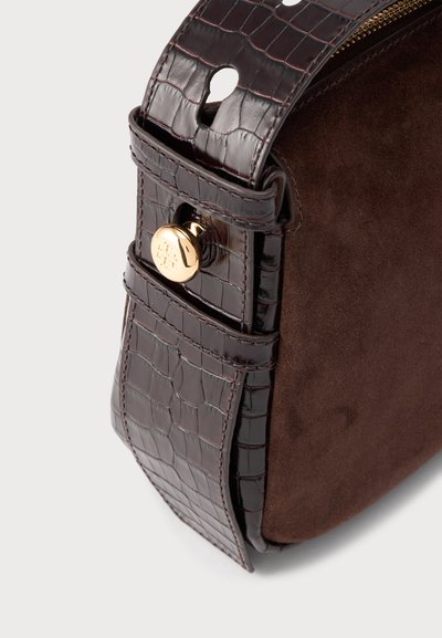 Angle de sac à main en daim brun foncé et cuir à motif crocodile avec bouton-pression doré et fermeture éclair sur fond blanc.