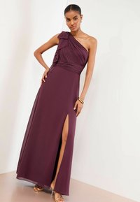 Burgundia ühe õlaga kleit volditud ja volangidega, millel on reie kõrge lõhike ning voolav tekstuur, sobitatud kuldsete kontsakingadega.