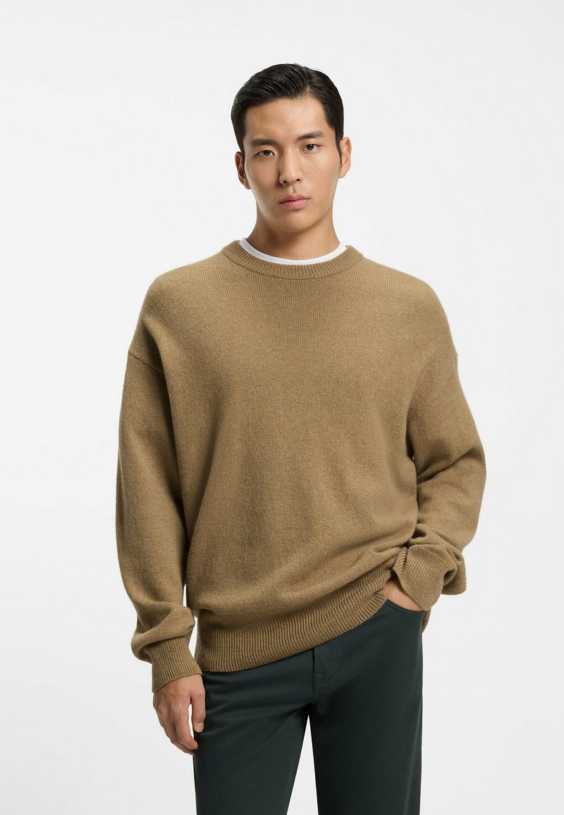 Maglione marrone chiaro a maglia con scollo rotondo e polsini a coste, abbinato a pantaloni verde scuro. La consistenza sembra morbida e leggermente elastica.