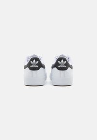 Baltas sporta apavi ar melniem papēža akcentiem, ar struktūru ādas virsu, apaļu purngali un klasisko Adidas logotipu aizmugurē.