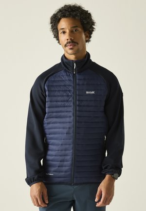 Mann trägt eine marineblaue, leichte gesteppte Jacke mit durchgehendem Reißverschluss und langen Ärmeln und steht vor einem einfarbigen hellen Hintergrund.