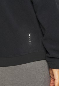 Giacca in pile nera con una texture morbida. Presenta un orlo aderente e un'etichetta con il logo 'RUSH' sul lato. Abbinata a pantaloni grigi.
