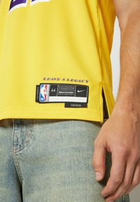 Maglia NBA gialla in tessuto a rete con dettagli viola, con un'etichetta che recita "LEAVE A LEGACY" e il marchio Nike all'orlo.