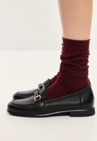 Zwarte leren loafers met een kleine hak, voorzien van een zilveren gespdetail. Draagt met bordeauxrood gestructureerde enkelkousen.