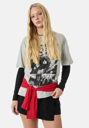 Young woman with long blonde hair wearing a grey graphic t-shirt over black long sleeves, black skirt, and a red sweater tied around her waist.