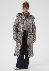 Donna in piedi che indossa un lungo piumino leopardato sopra pantaloni larghi scuri e scarpe da ginnastica bianche su uno sfondo semplice.