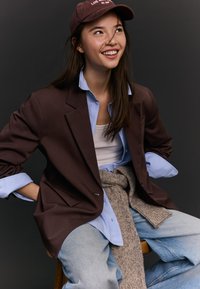 Mlada ženska, oblečena v rjav blazer, modro srajco, kavbojke in rjavo kapo, se smehlja in sedi ob temnem ozadju.