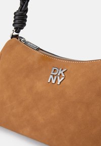 Barna műsuede válltáska ezüst DKNY logóval, fekete fonott fogantyúval és cipzáros záródással a tetején. Simaság jellemzi.