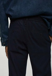 Černé kalhoty s elastickým pasem, s dvěma zadními kapsami na knoflíky, vystavené na pozadí džínové košile.