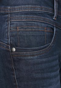 Tummansiniset farkut, joissa on kontrastoivaa oranssia tuskausta, pieni hopeinen nappi ja takataskun muotoilu. Sujuva pinta.