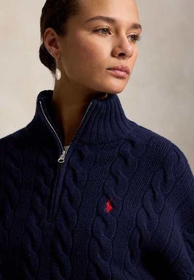 Nő sötétkék fonott mintás cipzáras pulóverben, piros hímzett logóval és egy lógó gyöngy fülbevalóval, oldalra nézve.