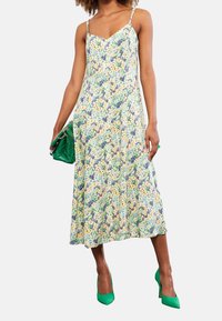 Florales Midikleid mit dünnen Trägern, mit einem mehrfarbigen Muster in Blau, Gelb und Grün. Kombiniert mit grünen High Heels und einer Clutch.