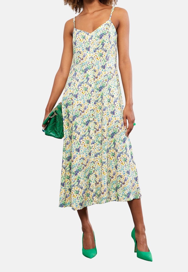 Florales Midikleid mit dünnen Trägern, mit einem mehrfarbigen Muster in Blau, Gelb und Grün. Kombiniert mit grünen High Heels und einer Clutch.