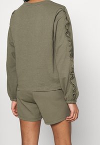 Felpa verde oliva con dettagli arricciati lungo le maniche, abbinata a pantaloni corti coordinati. Materiale in cotone, vestibilità comoda e maniche a sbuffo.